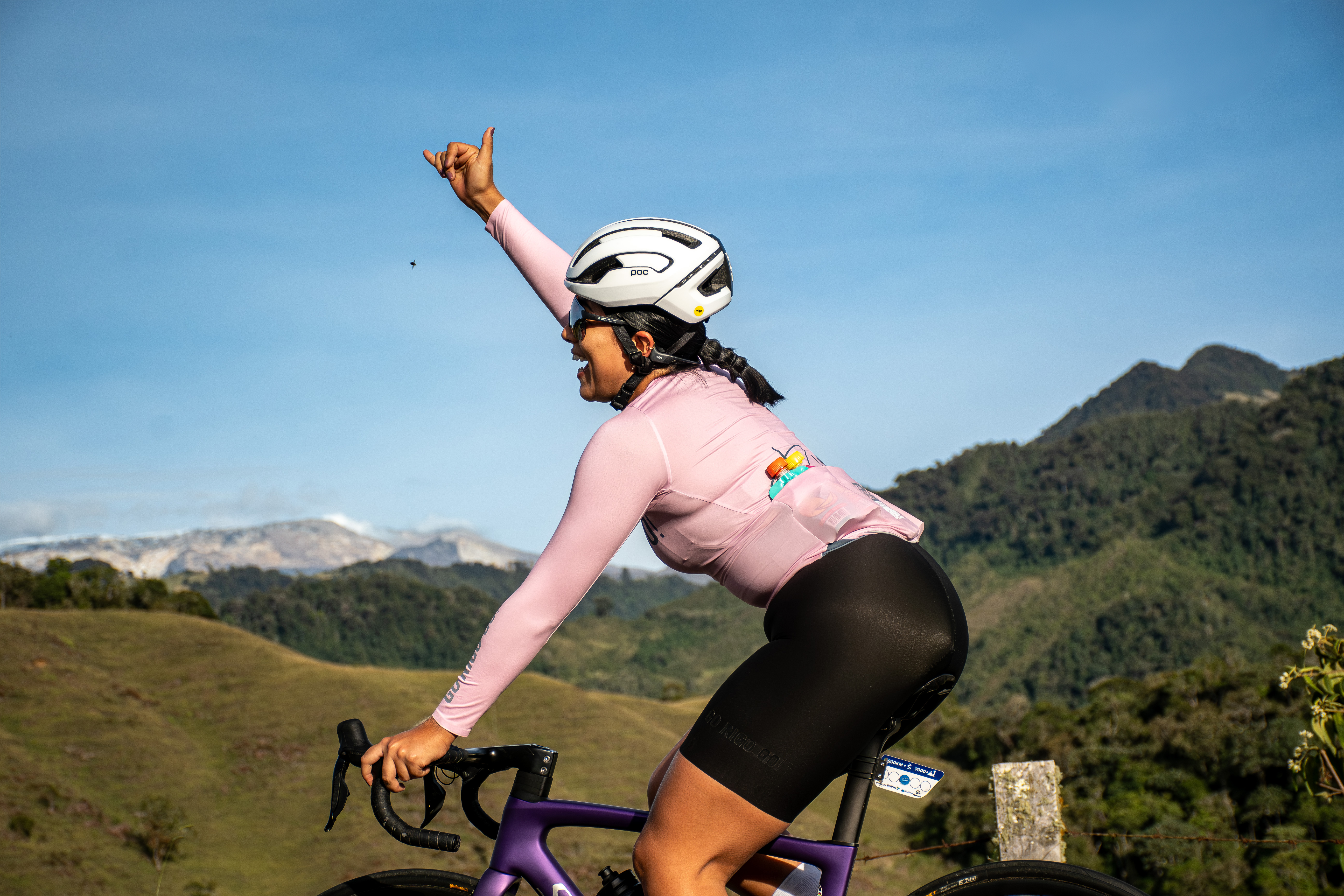 Female cyclist celebrating with a hand gesture while riding a road bike through the mountain scenery of Colombia.