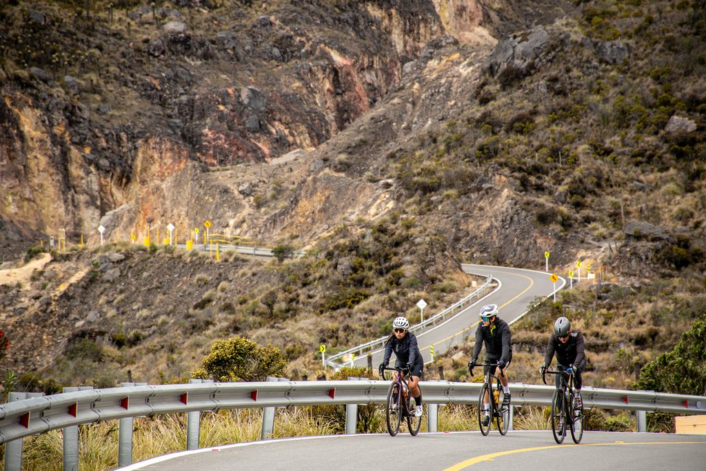Three cyclists in full gear and helmets riding up a winding, scenic mountain road during a Beyond The Ride all-inclusive cycling tour to Alto de Letras, Colombia.