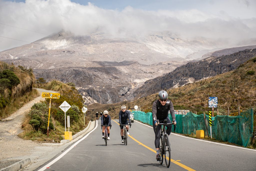Three cyclists in full gear riding up a paved mountain road through a paramo ecosystem towards a large, cloud-capped peak during a Beyond The Ride cycling tour in Colombia.