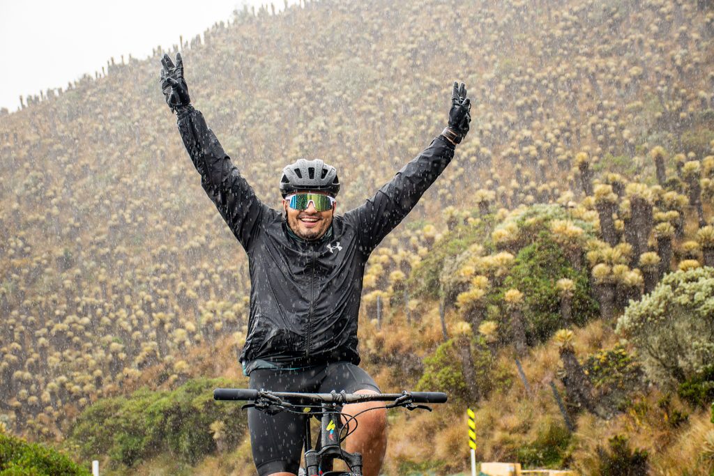 Happy cyclist wearing black rain jacket, sunglasses, and helmet celebrating with arms raised in the air while riding through light rain in a paramo landscape covered with frailejones during a Beyond The Ride cycling tour