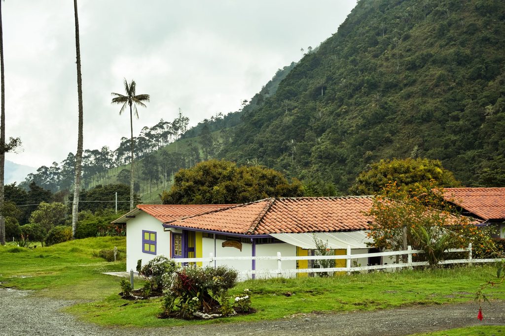 Traditional white coffee hacienda with red tile roof surrounded by green mountains and wax palms in Colombia's Eje Cafetero.