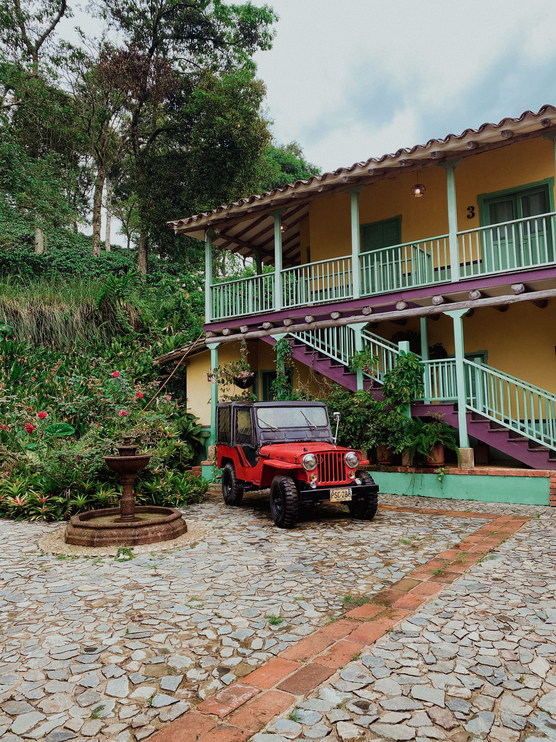 Red vintage Jeep Willys parked in the cobblestone courtyard of a traditional colonial coffee hacienda in Colombia's Eje Cafetero.